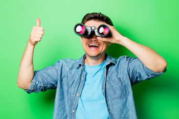 Ein lachender junger Mann schaut durch ein Fernglas und streckt dabei seinen Daumen nach oben.