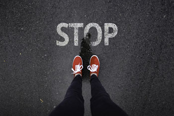 Aus der Vogelperspektive stehen Beine mit roten Schuhen vor einem, auf die Straße gemalten, STOP-Schriftzug.